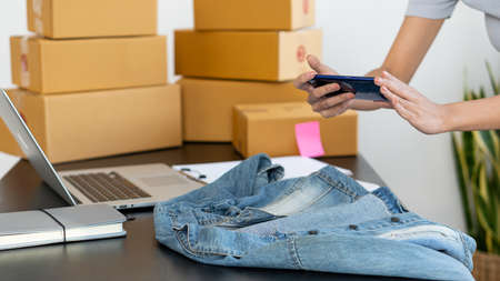 Online business, a young woman with a cellphone, take a picture of the clothes on the table, with a laptop and a box.の写真素材