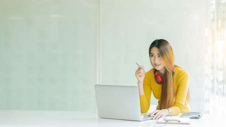 A young Asian woman who wears headphones on her neck is working on a laptop, focuses on students...