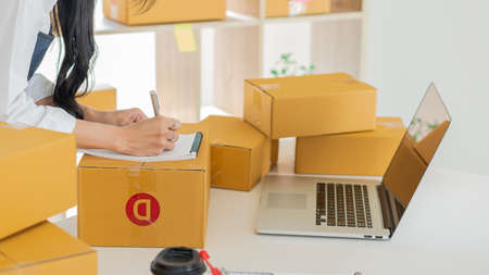 Young woman small business owner startups write address on cardboard box at work SME small business...