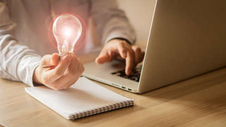 A business man wearing a gray shirt is sitting in front of a laptop holding a light bulb in his hand working technology ideas and ideasの写真素材