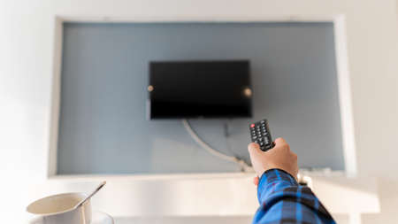 A young man with a remote control and a coffee mug is watching TV in the room.の写真素材