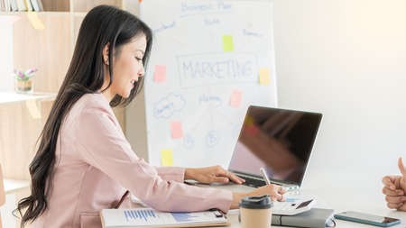 A beautiful Asian businesswoman works with documents, graphs, analyzes, and uses a laptop to work...