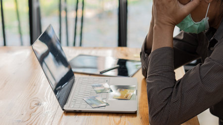 A man with his hand over his face worried about credit card debt with a laptop on his deskの写真素材