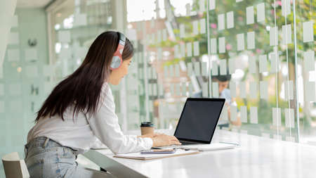 Young Asian students wearing headphones using their laptop in a coffee shop, writing notes on...