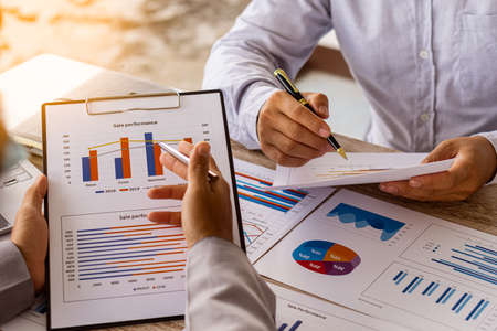 Close-up of two businessmen with pens pointing to graphs while chatting on wooden table with laptop,...