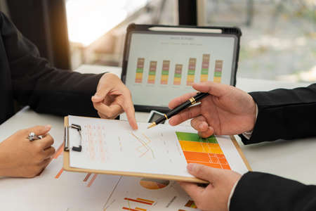 Close-up of two businessmen with pens pointing to graphs while chatting on wooden table with laptop,...