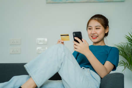 Asian woman sitting on sofa holding credit card and shopping from smartphone online. at home for online banking use mobile online shoppingの写真素材