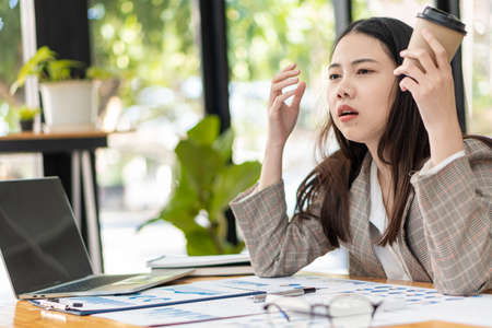 Asian businesswoman with a cup of coffee and drowsy or snoozing at the desk, an accountant who is tired of files in the office financial documents. need more restの写真素材