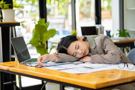 Asian businesswoman with a cup of coffee and drowsy or snoozing at the desk, an accountant who is tired of files in the office financial documents. need more restの写真素材