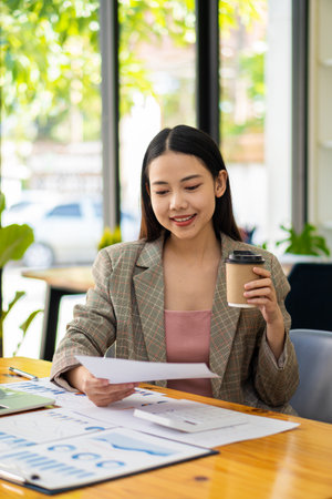 Smiling beautiful Asian businesswoman holding coffee cup and laptop working on graph papers at financial accounting concept officeの写真素材