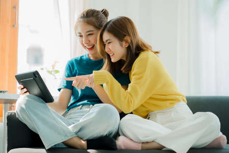 Two Asian women sit on gray sofas holding tablets while watching movies and listening to music on social media in their home living room.の写真素材