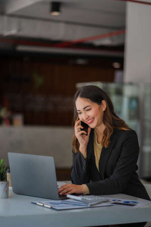Asian businesswoman or accountant working with laptop and mobile phone. Smile and be happy while sitting at your desk with your financial documents. Vertical modern shape office calculatorの写真素材