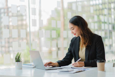Smiling beautiful Asian businesswoman sitting with laptop and computer working on paperwork make an account analysis report real estate investment information financial and tax system conceptsの写真素材
