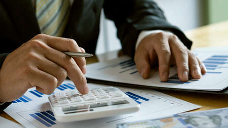 At the accounting desk, an accountant presses a calculator to analyze investment charts to generate...