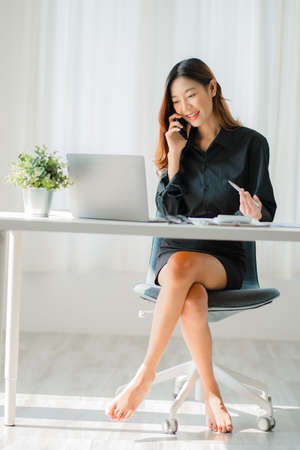 Asian businesswoman holding a smartphone analyzing a report pointing to a graph with a pen, laptop, and calculator. In workstation, accountant, online marketing concept, e-commerceの写真素材