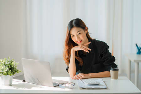 Asian businesswoman analyzing a report pointing to a graph with a pen, laptop, and calculator. In workstation, accountant, online marketing concept, e-commerce, landscape image.の写真素材