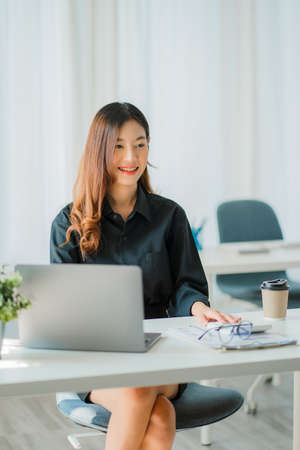 Asian businesswoman analyzing a report pointing to a graph with a pen, laptop, and calculator. In the online marketing concept of workstation accountant's vertical e-commerce landsの写真素材