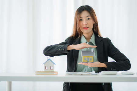 Real Estate Investment and Insurance Ideas Smiling Asian woman working with scale model house and house keys, contract documents in sales office.の写真素材
