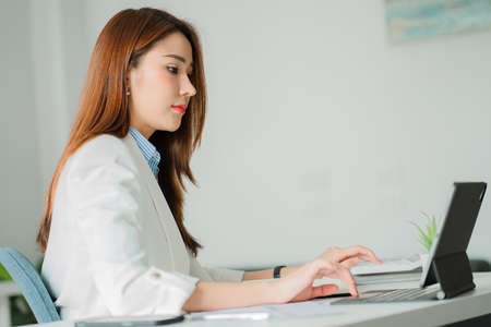 Asian woman sitting at a desk in a modern office with a tablet and taking notes online chatting with...