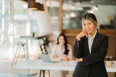 A business woman in a black suit looks good standing at her desk in a modern office. and colleagues working behind the scenesの写真素材