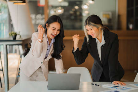 Two Asian business women rejoice at their success in business and raise their hands in front of their laptop computers. Colleagues rejoiced at the victory of the group.の写真素材