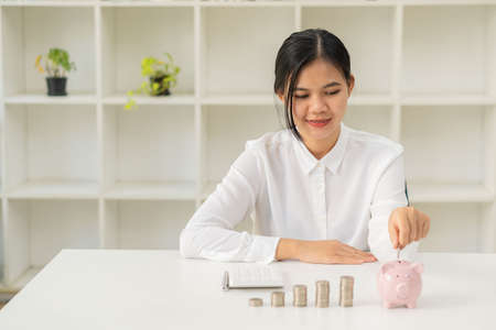 Smiling Asian woman holding a coin in a jar with piles of coins on the table to manage money and make a savings plan. future growth personal economy business and financial conceptsの写真素材