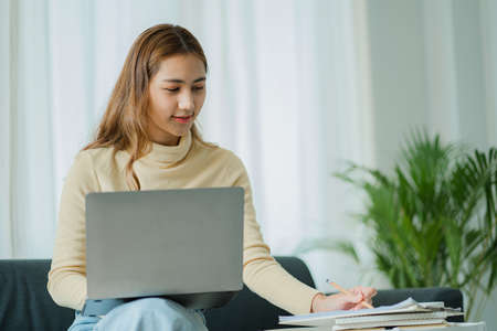 A freelance Asian woman working on a laptop checks social media while sitting on the sofa relaxing in her living room at home. concept of lifestyle for womenの写真素材