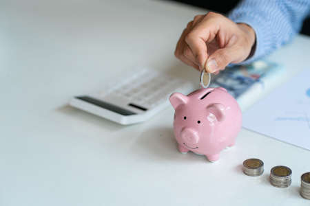 A businessman puts a coin in a jar and a calculator sits next to it. It is a concept in savings and...