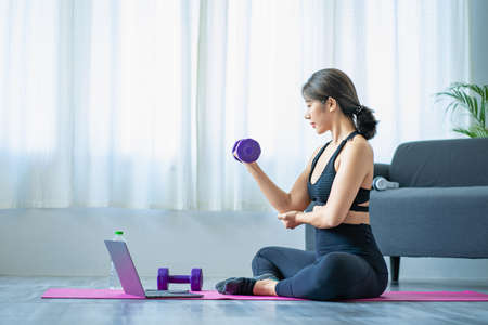 Smiling Asian woman doing shoulder stretching yoga online class from laptop at home in living room. Self-isolation and exercise at home with a sofa and laptop next to it.の写真素材