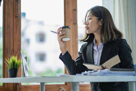 Asian businesswoman working in office with papers and holding pen using laptop sitting on chair at...