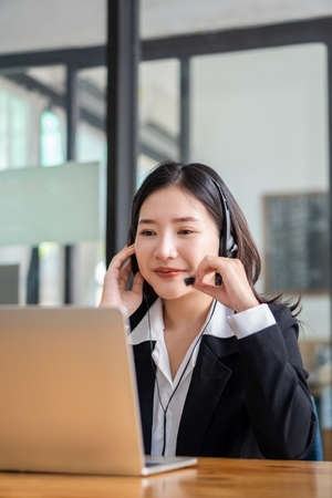 Charming and smiling Asian female customer service worker wearing headset in online meeting office...