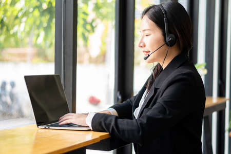 Charming and smiling Asian female customer service worker wearing headset in online meeting office look at laptop computer screen, work and social distancing concept, financial busの写真素材