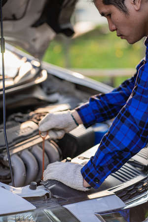 An Asian man looks under the hood of a broken car and checks the oil level on the side of the road.