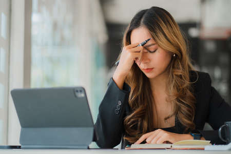 A stressed Asian accountant sitting in front of her laptop screen with her eyes closed at the desk....