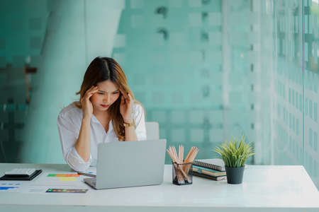 Asian businesswoman having a headache during work analyzing financial graphs with laptop at office...