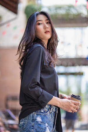 A beautiful Asian woman holding a cup of coffee in a black shirt is smiling happily and standing in...