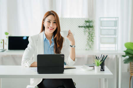 Beautiful smiling Asian businesswoman working using a tablet at the office while sitting in a modern...
