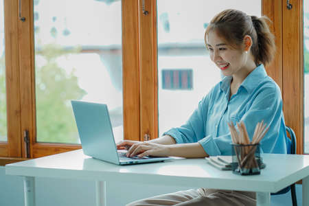 Smiling Asian woman working with laptop computer at her desk in modern home office. calculating financial expenses Make a note on the graph paper data sheet.の写真素材