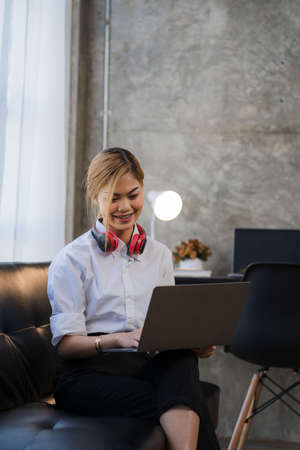 Smiling beautiful Asian woman working on a laptop sitting on the sofa at home online learning...