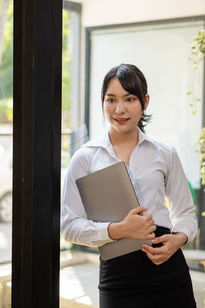 A young accountant stands with her laptop next to a glass wall in her office.