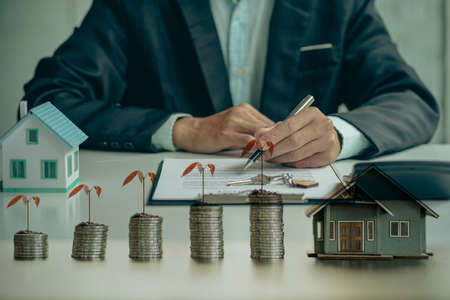 businessman holding pen with papers and wooden house next to pile of coins with tree above real estate business growth ideaの写真素材