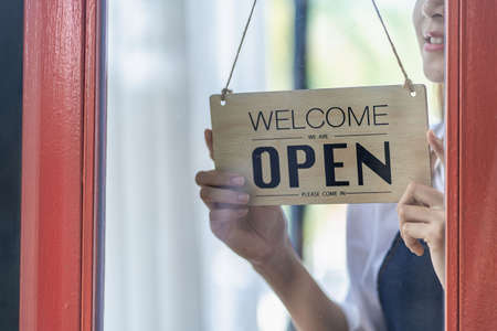A female barista holding a welcome sign in front of a cafe in front of the shop while standing....