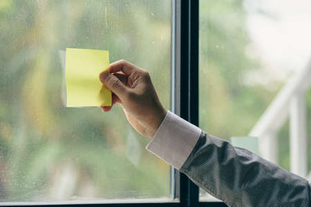 Young businessman hand holding a post-it colored paper pasted on glass wallの写真素材