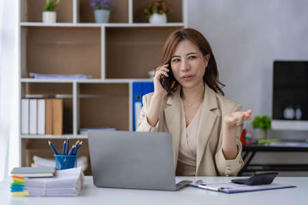Attractive Asian businesswoman talking on mobile phone in home office while analyzing monthly schedule in financial accounting concept laptop.の写真素材