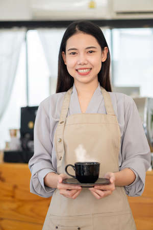 A young Asian barista holds a cup and stands in front of a counter in a coffee shop looking at the camera and smiling.の写真素材