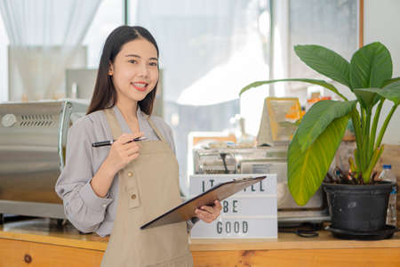 A young Asian barista holding a menu folder stands in front of a coffee shop counter, looks at the...