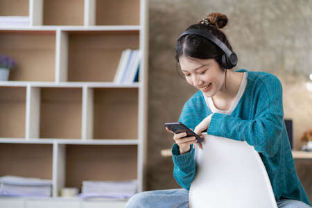 Cute Asian woman smiling using headset looking at smartphone screen listening to music on sofa at home.の写真素材