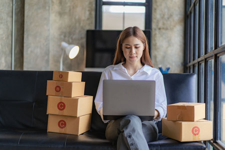 Asian woman sitting on sofa at home selling online with box and laptop receiving messages from customers sme business ideaの写真素材