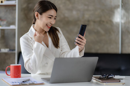 Attractive young Asian business woman holding smartphone working on laptop to analyze financial graphs on office tableの写真素材