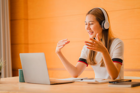 Attractive Asian college students wear headphones to watch laptop screens, study online or attend online meetings at home. women learn languages, listen to lecturesの写真素材